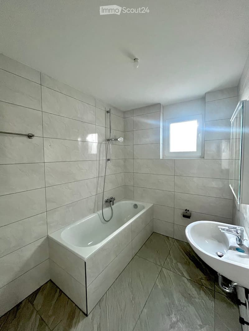 Modern bathroom with a bathtub, sink, and showerhead, featuring light grey tiles and a window for natural light.