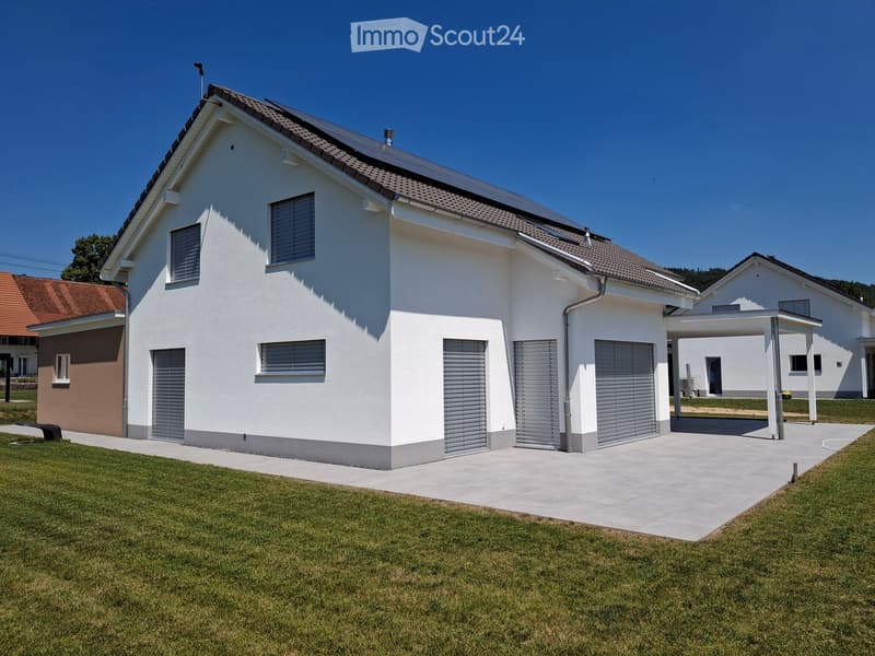 Modern two-story house with white exterior, grey shutters, and a covered driveway. The house features a garage and is surrounded by a well-maintained lawn.