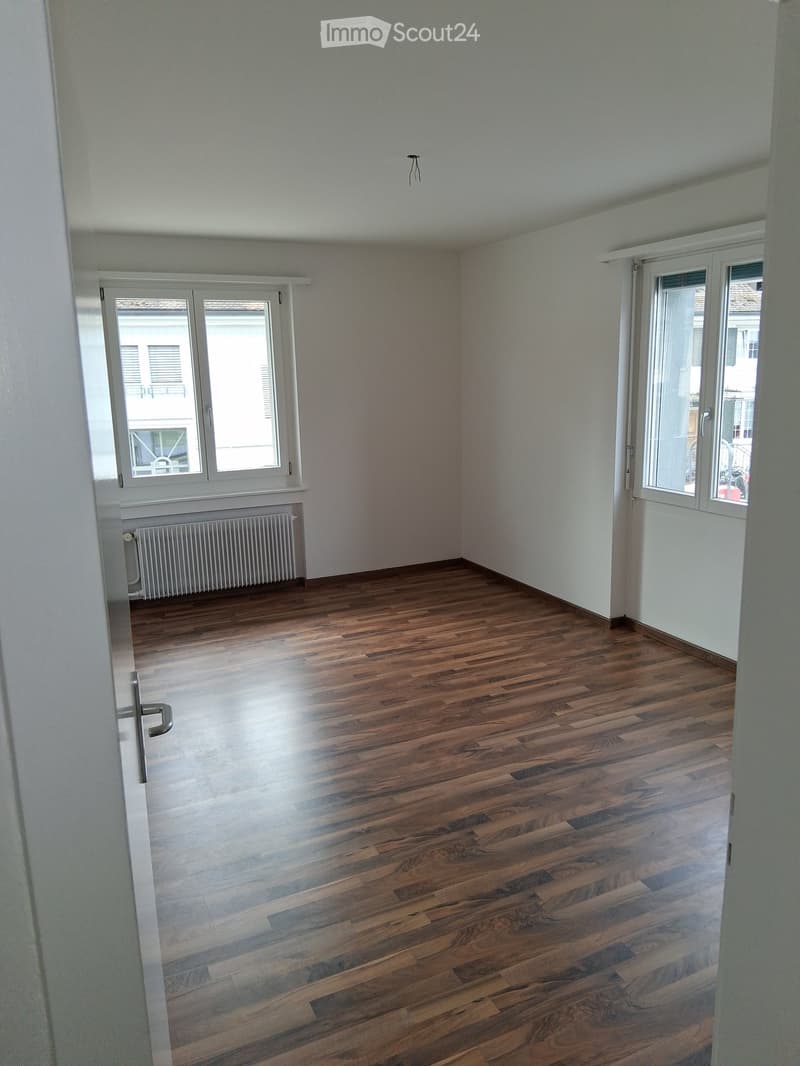Empty room with wooden flooring, white walls, and large windows with white frames. A radiator is visible under one window, and a door is seen on the left side.