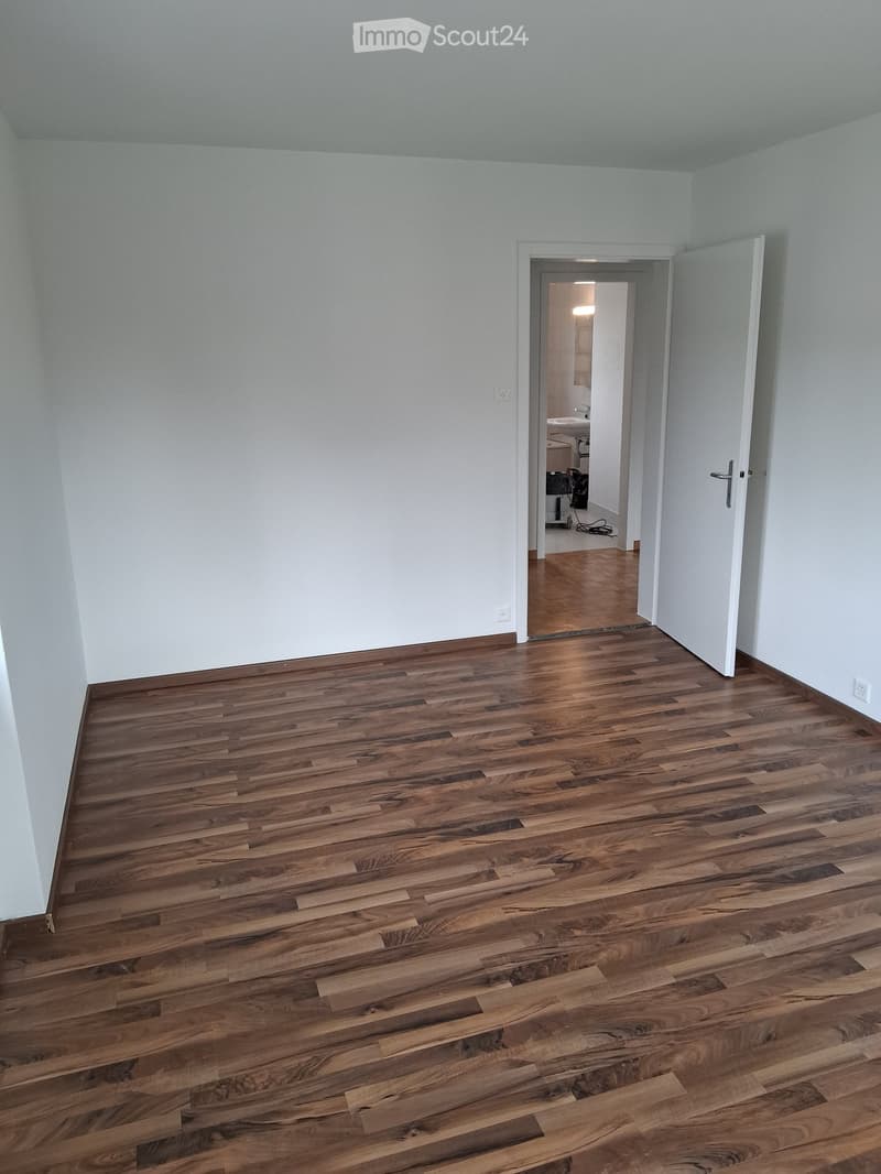 Empty room with wooden flooring and white walls, featuring an open doorway leading to another room with a visible sink and cabinets.