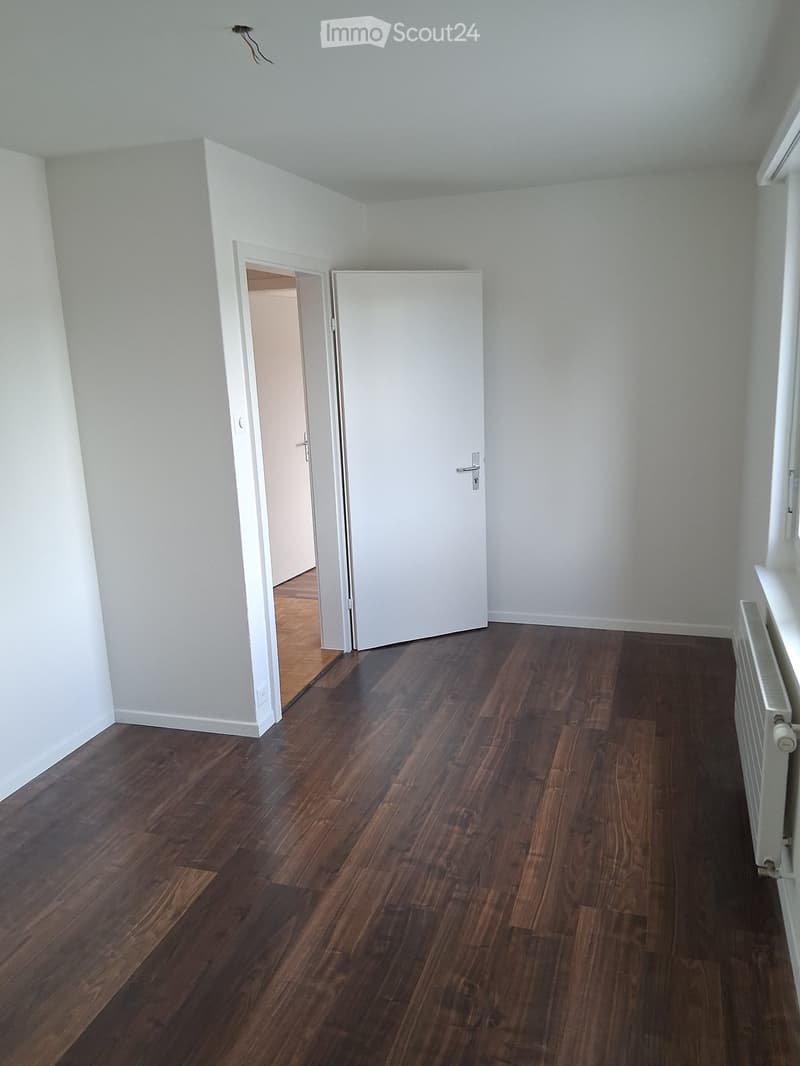 Empty room with wooden flooring, white walls, and a radiator on the right side. A white door is partially open, revealing another room with wooden flooring.