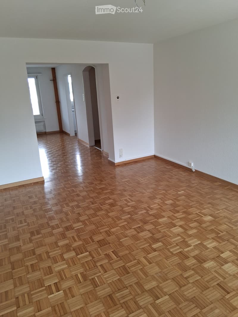 Empty room with wooden parquet flooring and white walls, featuring an arched doorway leading to another room.