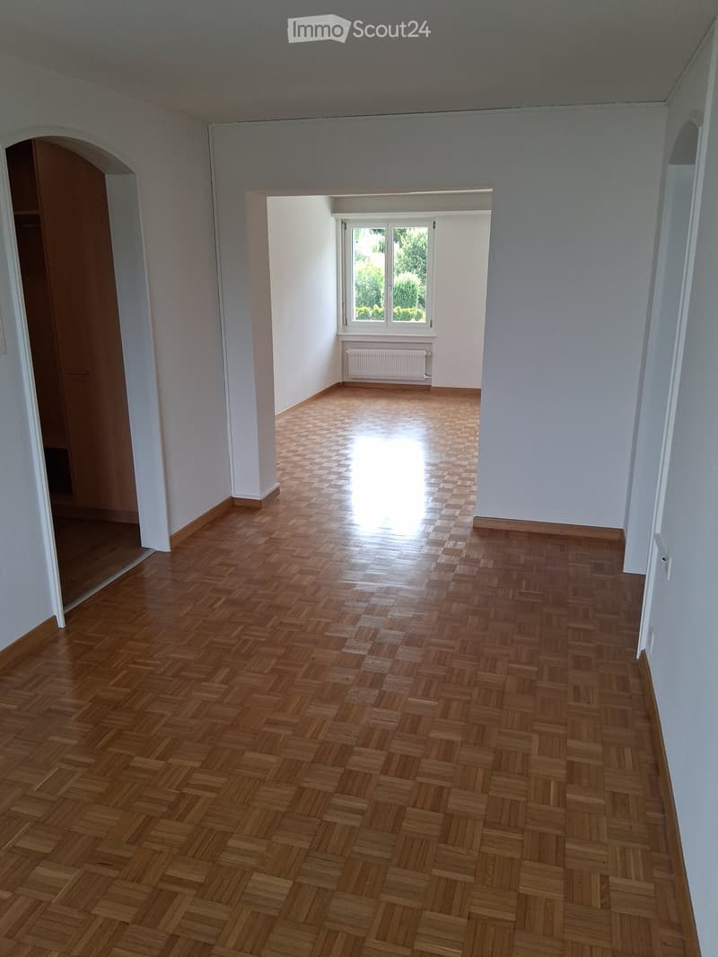 Empty room with wooden herringbone flooring, white walls, and a large window letting in natural light. The room features a radiator below the window and an open doorway leading to another area.