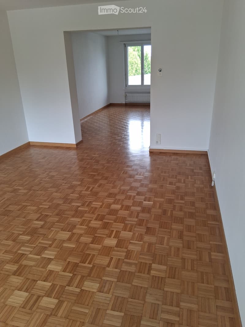Empty room with light-colored walls and a polished wooden parquet floor, featuring a visible doorway leading to another room with a window and radiator.