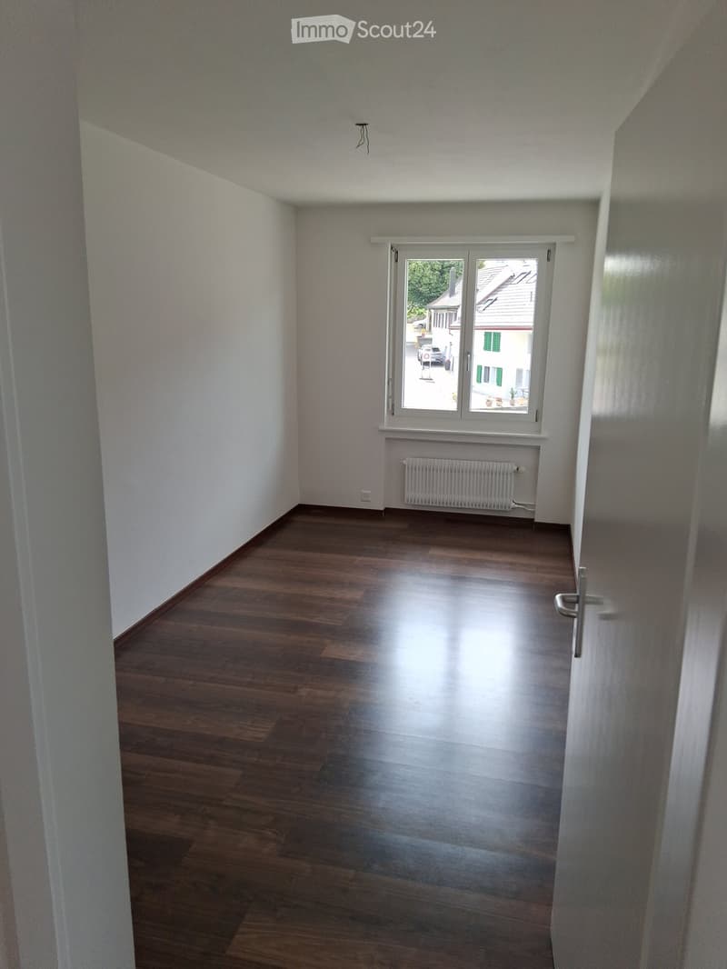 Empty room with wooden flooring, white walls, a window with a view of the street, and a radiator below the window.