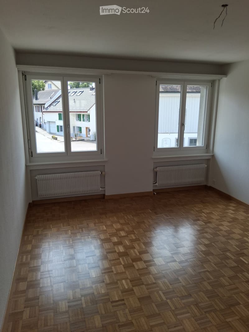 Empty room with wooden parquet flooring, large windows, radiators, and a sliding glass door leading to an outdoor area.