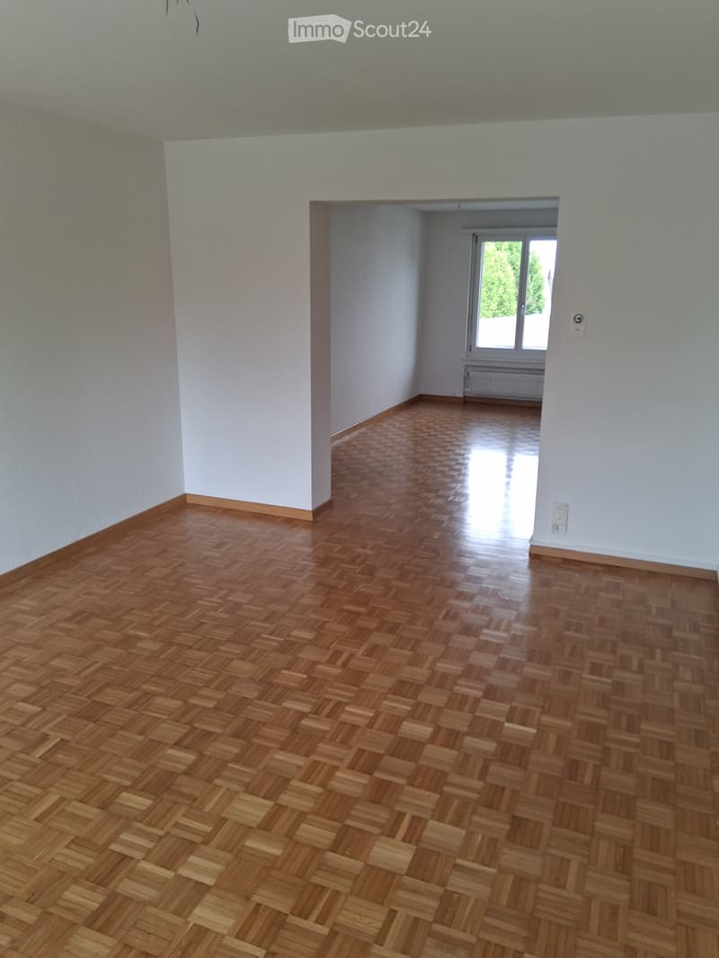 Empty room with wooden parquet flooring and white walls, featuring a doorway leading to another room with a window and radiator.
