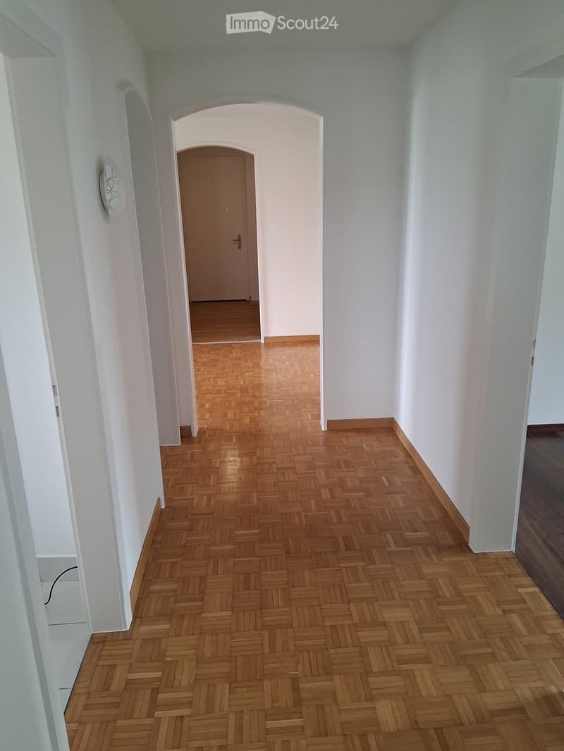 Empty hallway with wooden herringbone flooring, white walls, and a single door at the end.