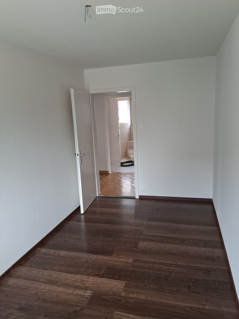 Empty hallway with wooden flooring and white walls, leading to a small bathroom with a toilet and a green cleaning spray bottle on the floor.