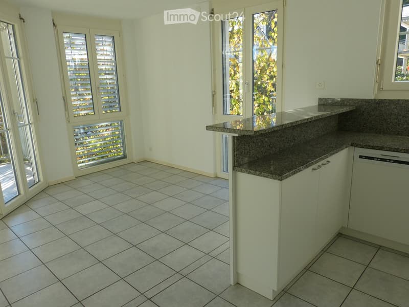 Empty kitchen with tiled floor, granite countertop, and white cabinets. Large windows with blinds and shutters provide natural light.