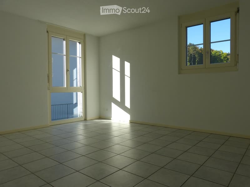 Empty room with tiled floor, white walls, and large windows allowing natural light. Features a sliding glass door leading to a balcony.
