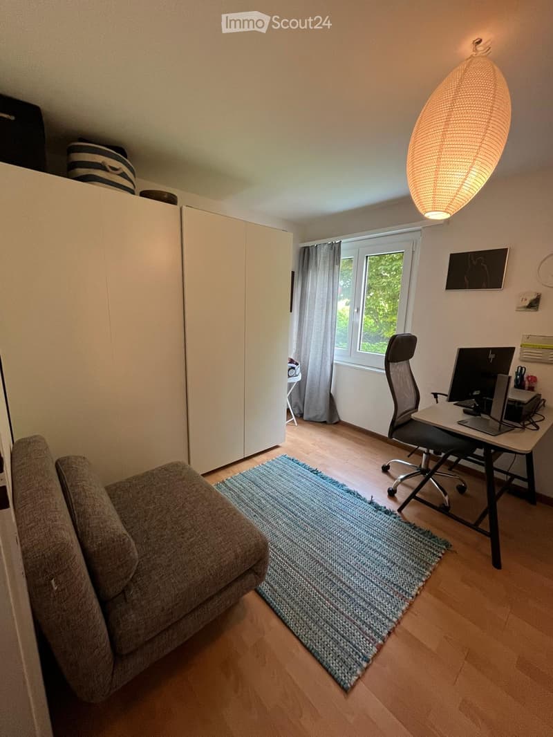 A cozy living and working space with a grey sofa, a modern desk setup, a large window with curtains, and a decorative lamp.