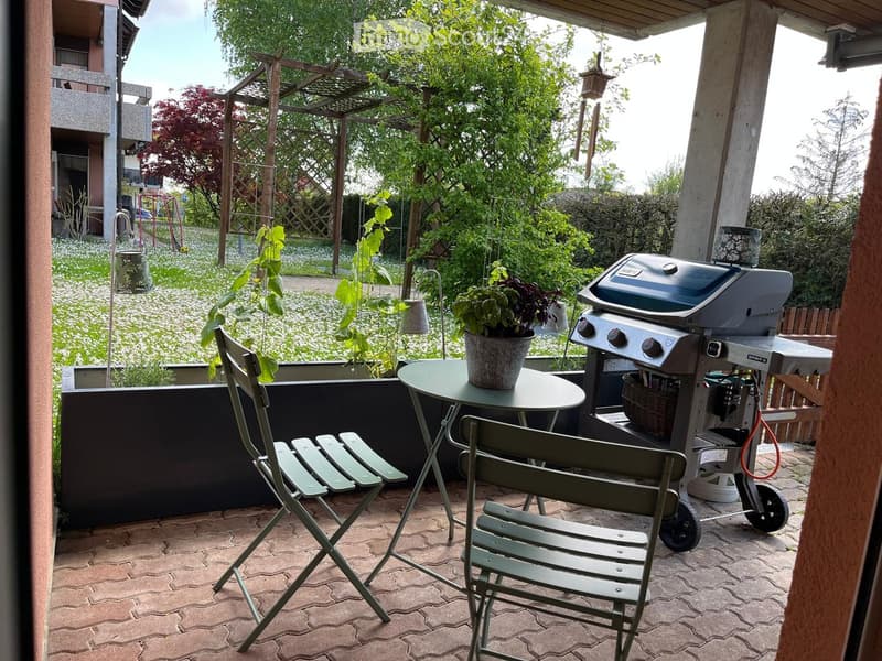 Outdoor patio with a round table, four folding chairs, a potted plant, and a grill. The area is surrounded by greenery and has a pergola overhead.