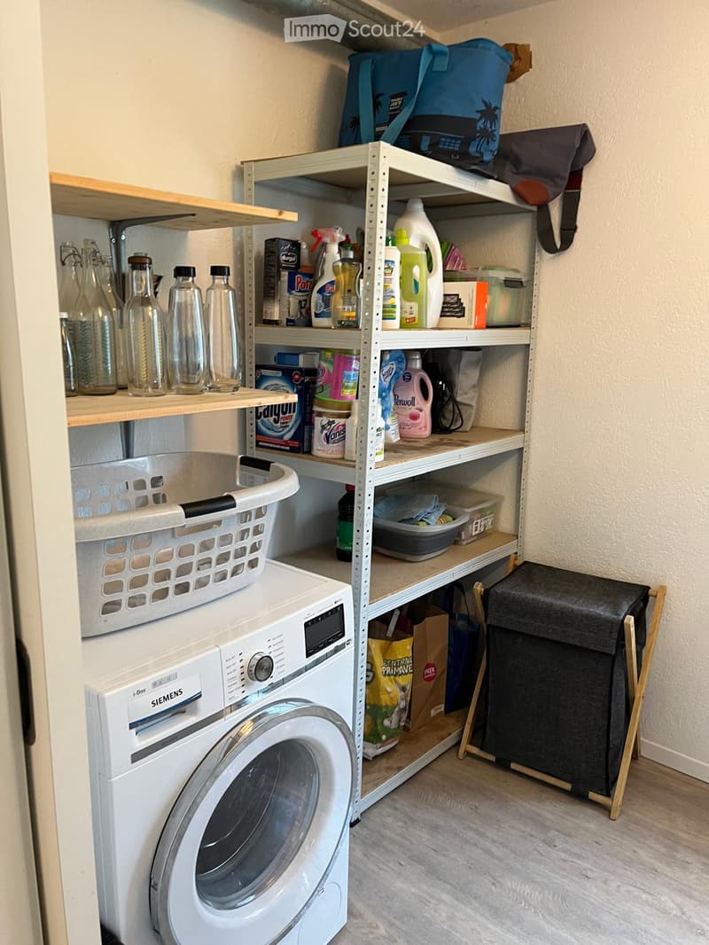 Laundry room with a washing machine, shelves holding cleaning supplies, a laundry basket, and a trash bin.
