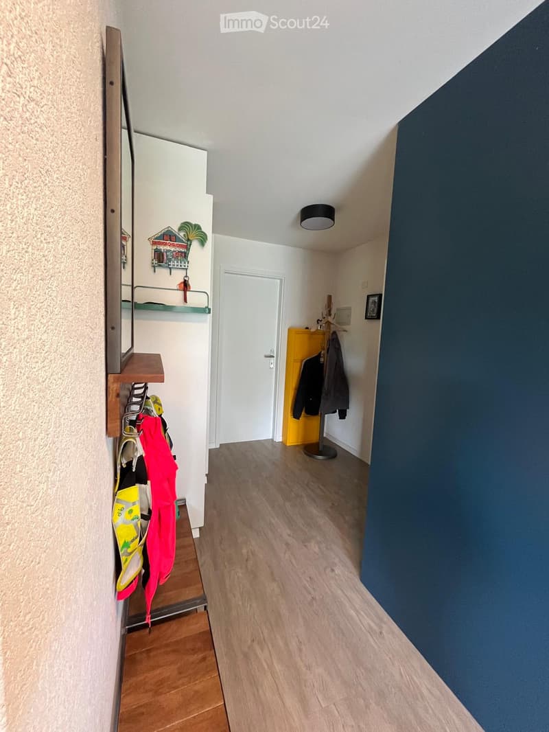 A narrow hallway with wooden flooring, white walls, and a blue accent wall. Features include a coat rack with jackets, a small shelf with shoes, and a coat stand with additional jackets.