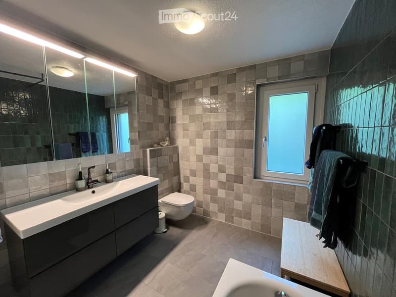 Modern bathroom with a large mirror, double sink, toilet, and bathtub. Features tiled walls and a window with frosted glass.
