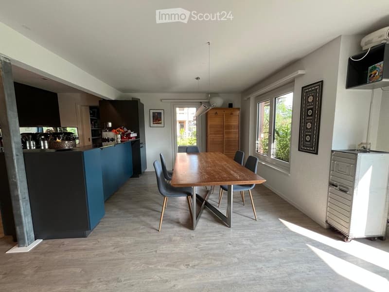 Open-plan kitchen and dining area with a wooden dining table, blue chairs, a kitchen counter with appliances, and a refrigerator. Large windows provide natural light.