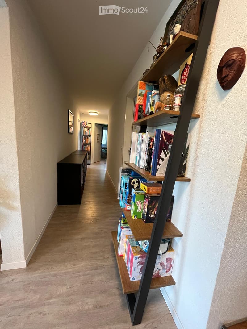 A well-lit hallway with wooden flooring, featuring a tall, leaning bookshelf filled with books and decorative items on the right side, and a dark wooden cabinet on the left side.
