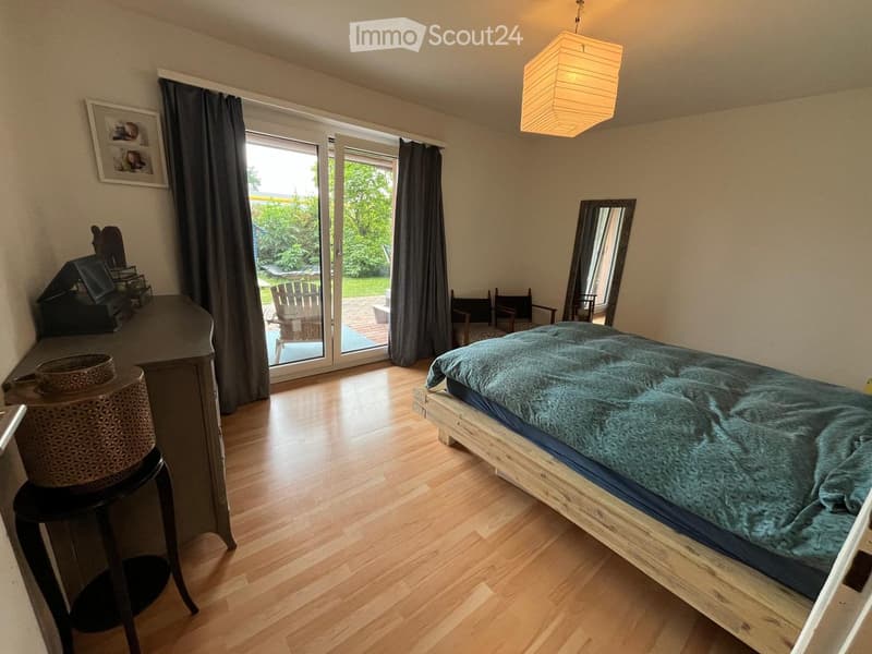 Cozy bedroom with wooden flooring, a large bed with a teal duvet, a mirror, a small table with a stool, and a view of a garden through large glass doors.