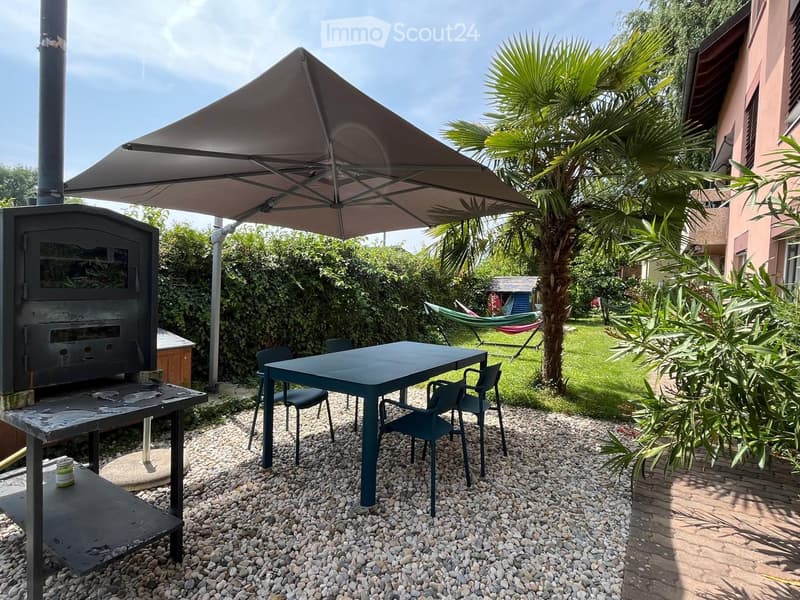 Garden with a table, chairs, a hammock, and a large umbrella, surrounded by greenery and a wooden stove.