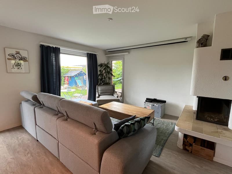 Modern living room with a grey sectional sofa, wooden coffee table, armchair, fireplace, and large windows overlooking a garden.