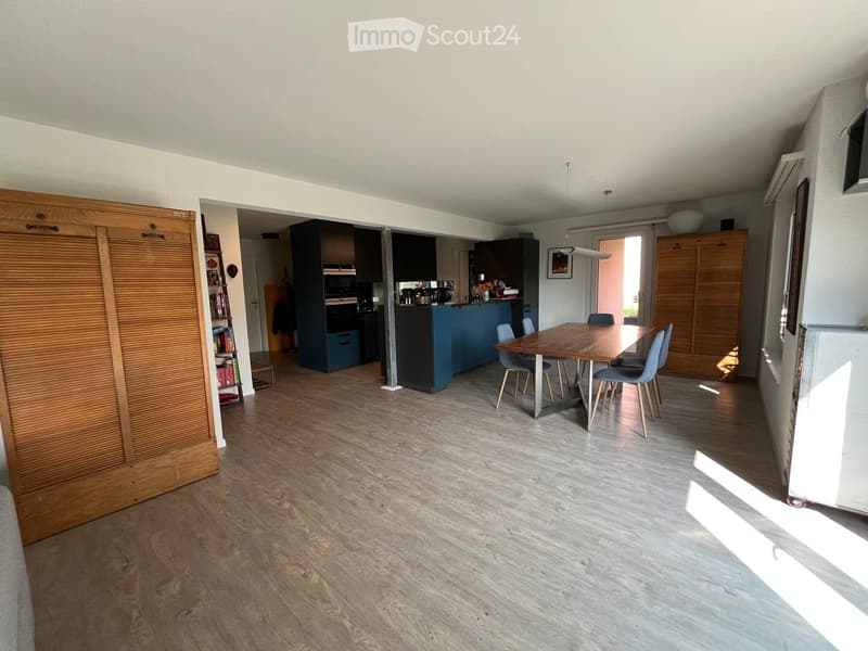 Open-plan living and dining area with wooden flooring, a dining table with chairs, a kitchen area with cabinets and appliances, and a bookshelf.
