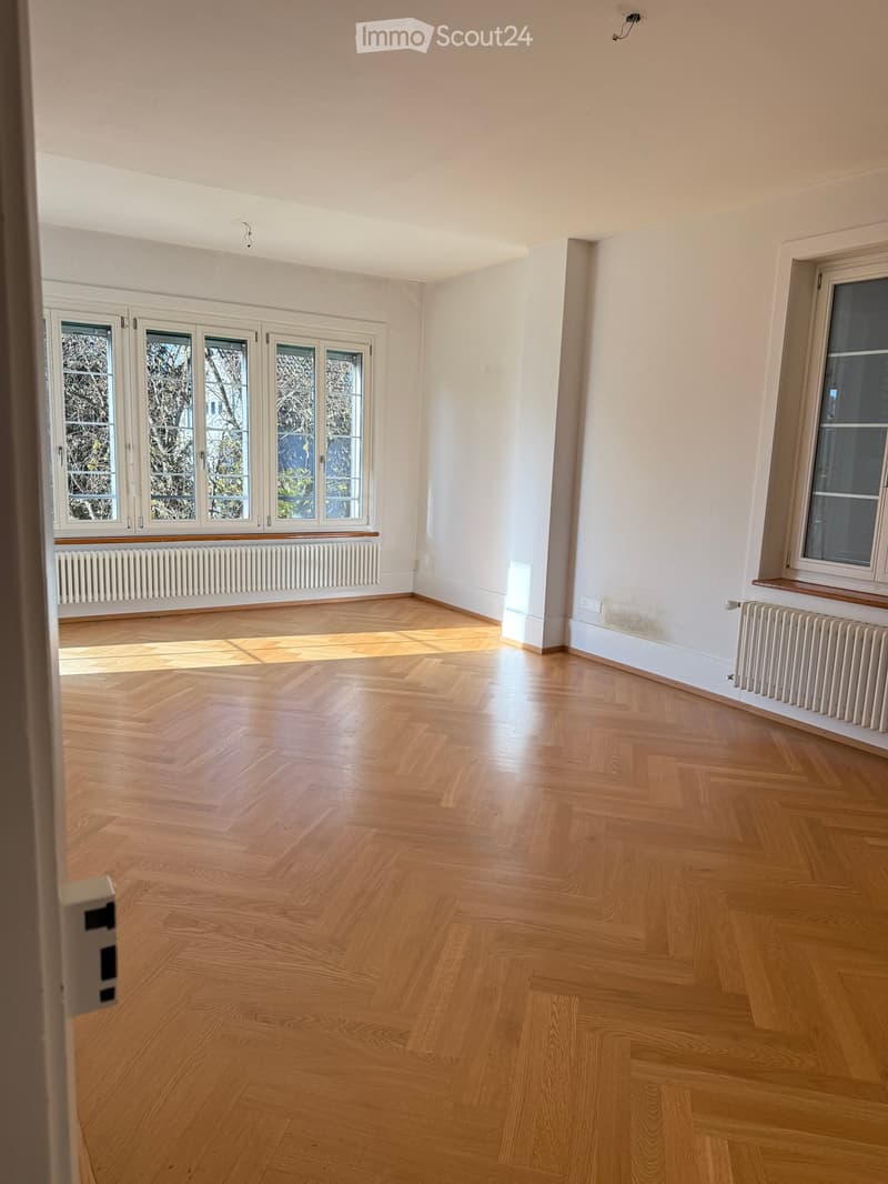 A spacious room with wooden parquet flooring and white walls. It features large windows with wooden frames that allow natural light. There are white radiators under the windows for heating. The room is empty.
