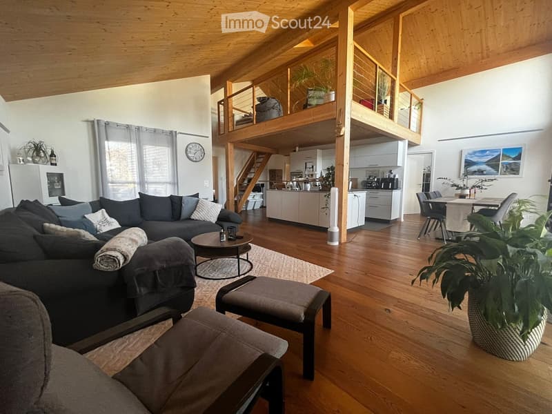 Open-concept living and dining area with wooden floors, a grey sectional sofa, coffee table, dining table, and kitchen area with white cabinets and appliances.