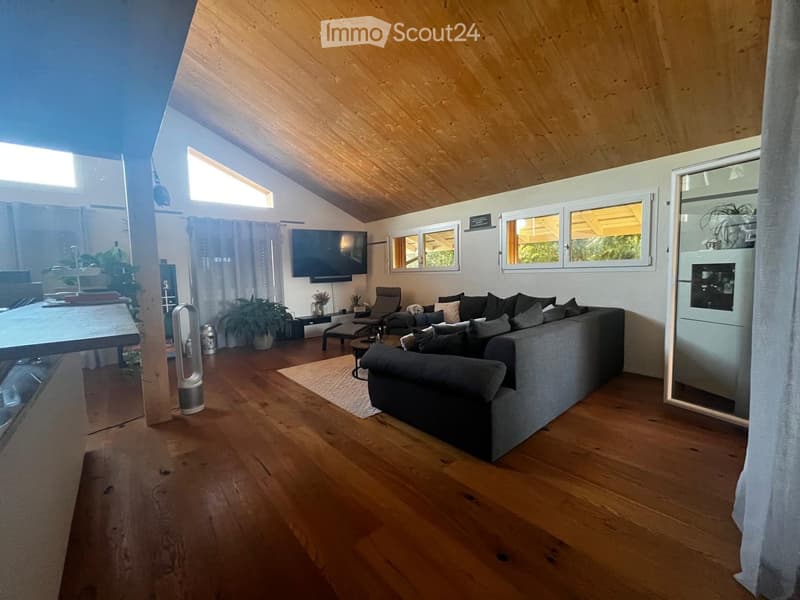 Modern living room with wooden flooring, large windows, a grey sectional sofa, coffee table, and a television mounted on the wall.