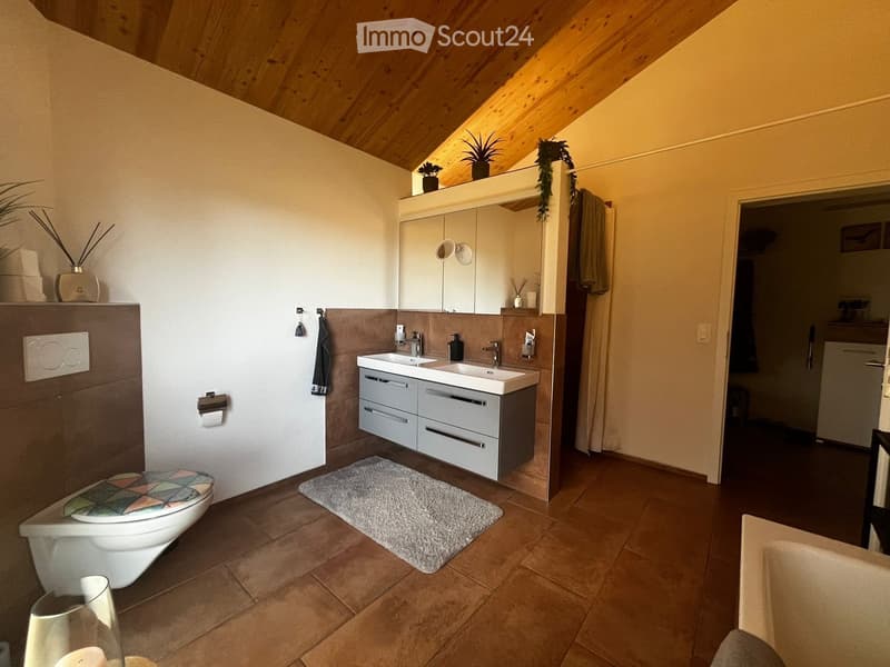 Modern bathroom with double sink, toilet, mirror, and built-in cabinets. Features a wooden slanted ceiling, tiled floor, and decorative plants on the vanity.