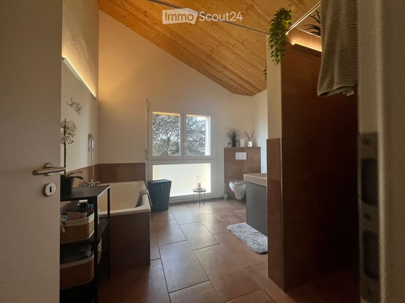 Modern bathroom with a bathtub, toilet, sink, and a small stool near the window. The room features a wooden slanted ceiling, tiled floor, and a small plant on the windowsill.