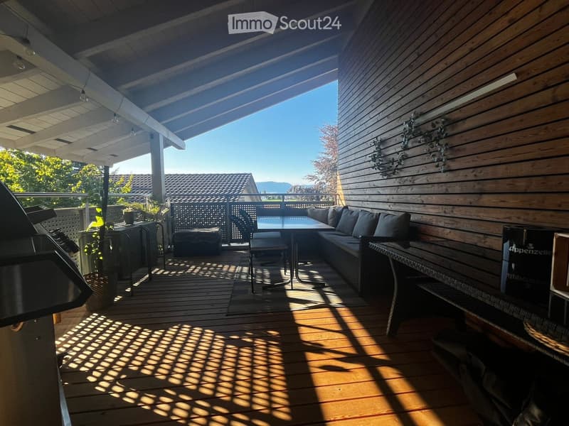 Wooden deck with a covered patio area, featuring a sofa, table, chairs, ottoman, and grill, with a view of the surrounding landscape.