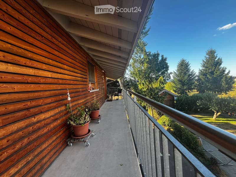 Balcony with wooden walls, metal railing, potted plants, and a view of a green landscape with trees.