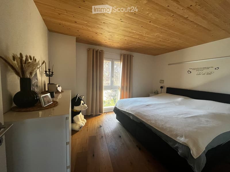 Cozy bedroom with wooden floor, slanted wooden ceiling, large window with curtains, bed with white linens, nightstand, decorative vase, candle holder, picture frame, and stuffed animal on floor.