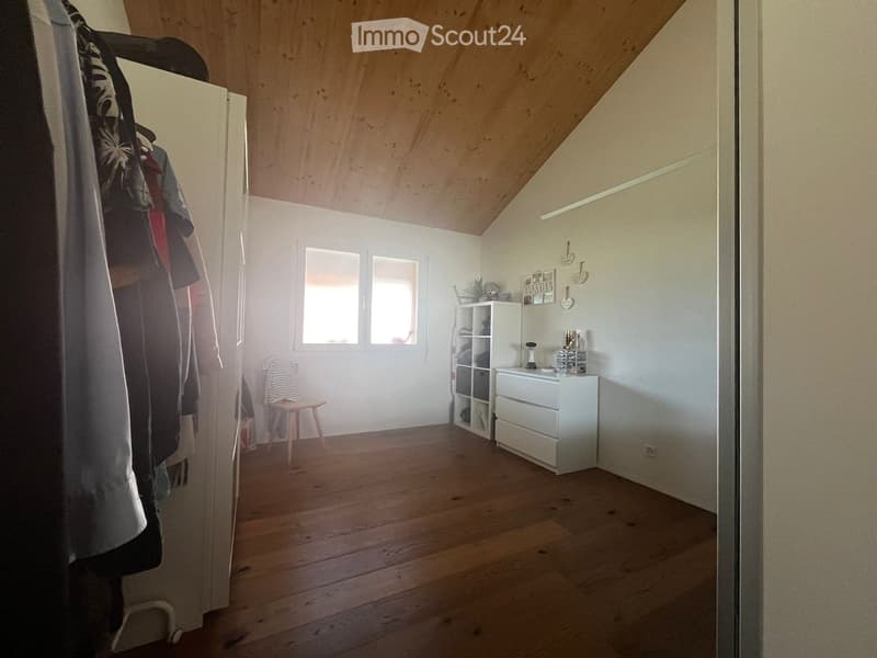 Bright, wooden-floored room with slanted ceiling, white walls, and large windows. Contains a small wooden stool, a white cabinet with drawers, a shelf, and a mirror on the wall.