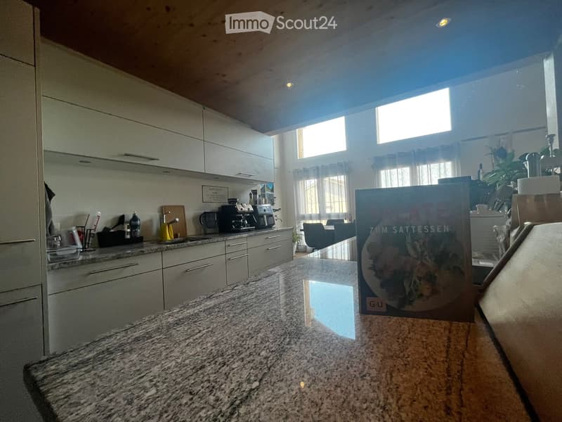 Modern kitchen with white cabinets, granite countertops, and a coffee machine on the counter. A book is placed on the counter, and a window allows natural light to enter.