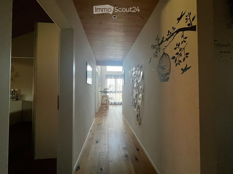 A narrow hallway with wooden flooring, light-colored walls, and a decorative mural featuring birds and a birdcage. A small plant and a small table are visible in the background.