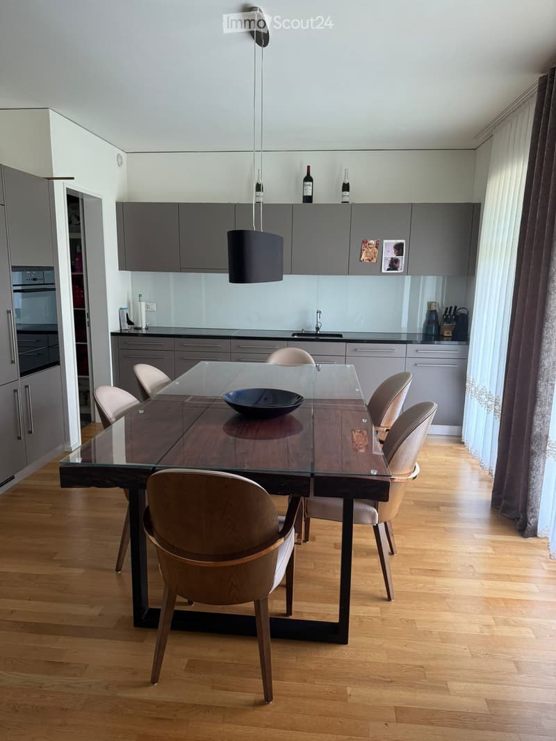 Modern kitchen with grey cabinets, a glass dining table, six chairs, a bowl on the table, and a hanging pendant light. The kitchen features a sleek design with a wooden floor and large windows with curtains.