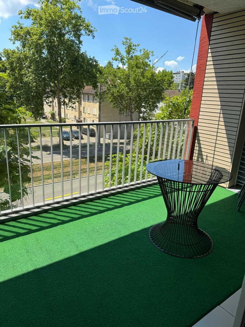 Balcony with green artificial turf, modern black metal table and chair, metal railing, and view of street and trees.