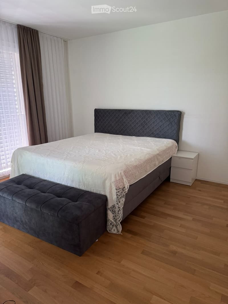 Modern bedroom with a large bed, quilted headboard, nightstand, and a grey bench at the foot of the bed. Wooden flooring and large windows with curtains.
