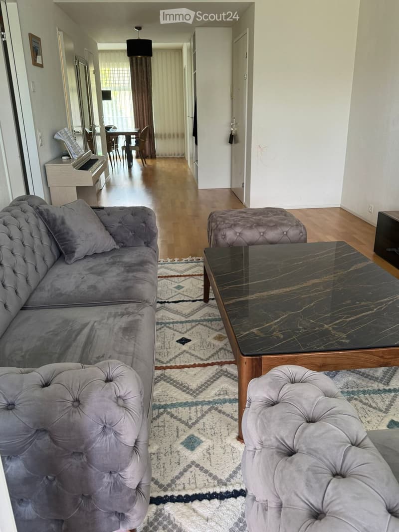 Living room with a grey tufted sofa, coffee table, ottoman, and a small piano in the background. Wooden flooring and patterned rug.