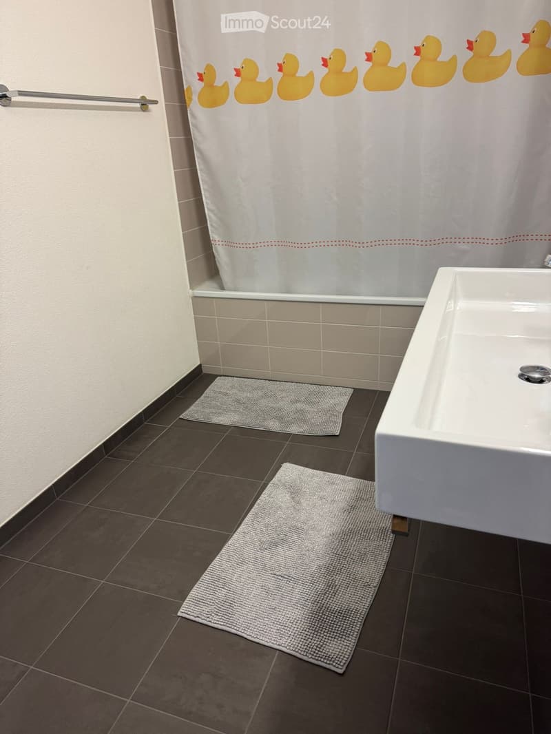 Modern bathroom with a bathtub, sink, and two grey rugs on dark tiled floor. Shower curtain features yellow rubber ducks.