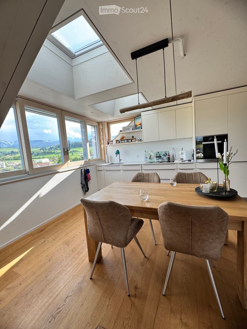 Modern kitchen and dining area with wooden flooring, large windows offering a scenic view, a dining table with four chairs, and white cabinets.