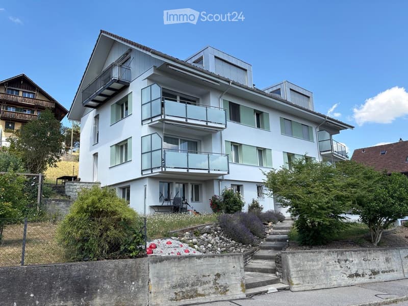 Modern two-story house with white exterior, multiple balconies, large windows, and landscaped garden with steps leading up to the entrance.