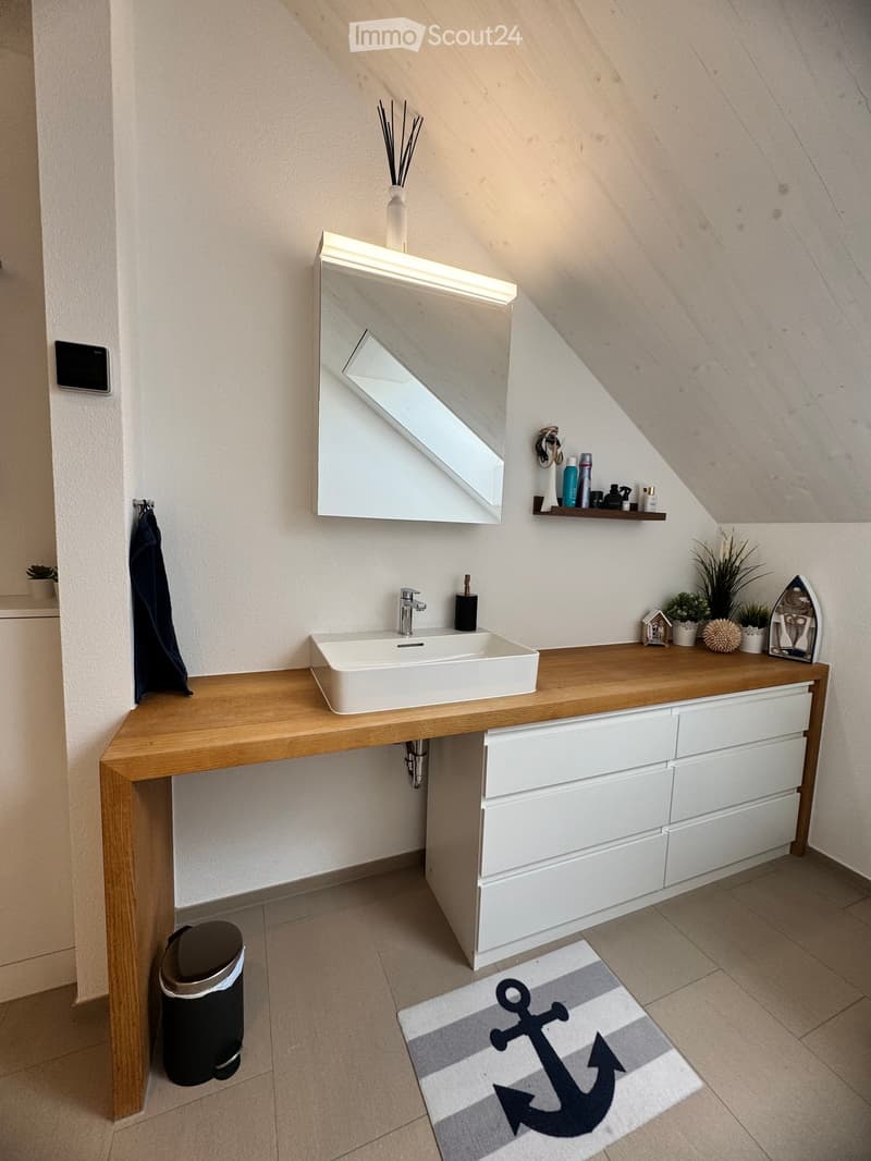 Modern bathroom with a wooden vanity, white sink, large mirror, and decorative items including a vase, plants, and a shelf with toiletries.