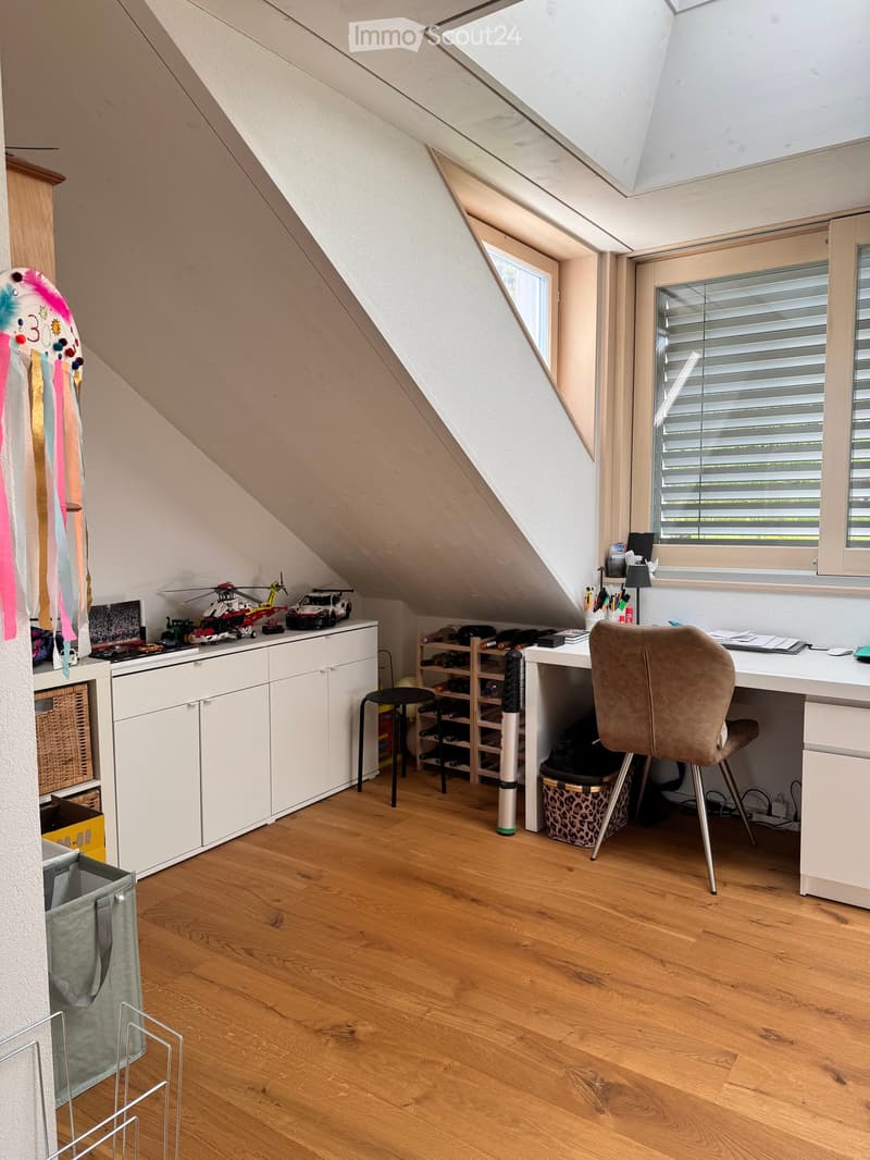 Attic room with wooden flooring, slanted ceiling, skylight, and large window. Contains a desk with a chair, a cabinet, a shelf, a stool, and various toys and items.
