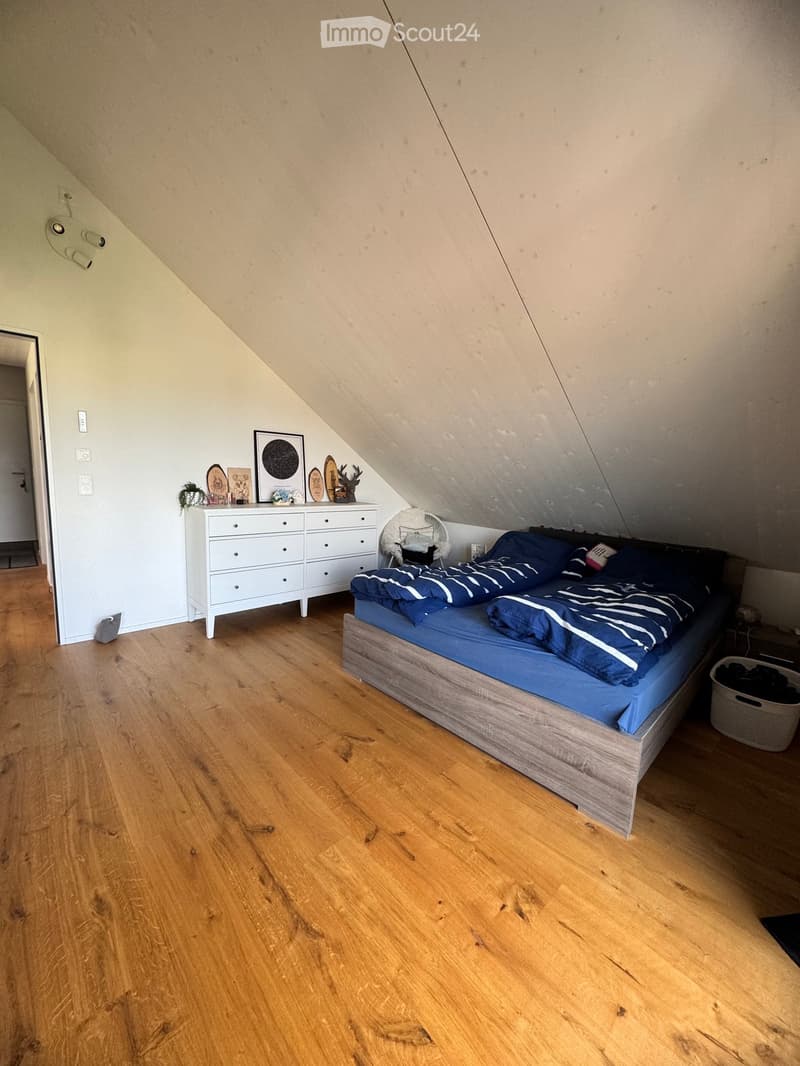 Cozy attic bedroom with wooden flooring, slanted ceilings, a bed with blue bedding, a white dresser, a fan, and decorative items on the dresser.