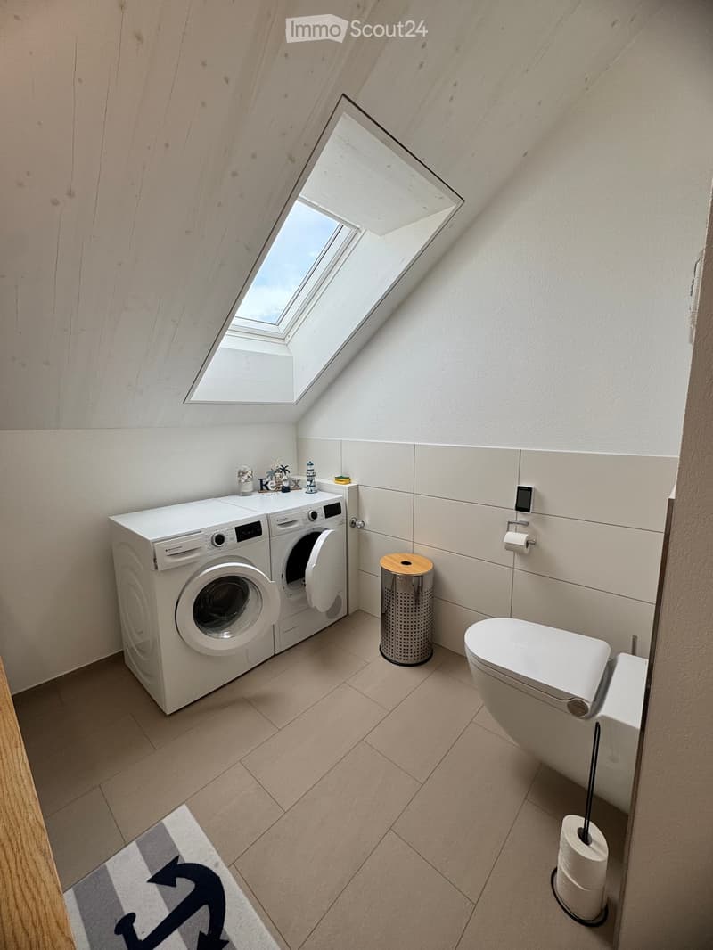 Modern bathroom with a skylight window, white washing machine, toilet, trash can, and decorative items on the washing machine.