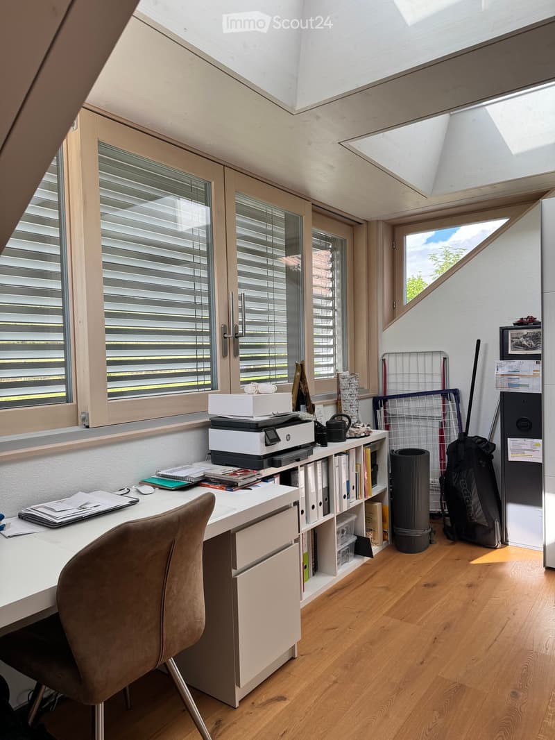 Bright, modern workspace with a white desk, ergonomic chair, printer, laptop, books, and filing cabinet. Large windows with blinds and a skylight provide ample natural light.