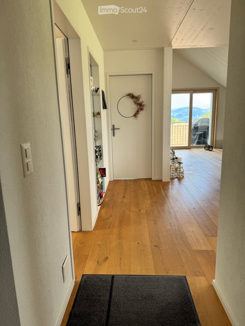 A well-lit hallway with wooden flooring, white walls, and a black doormat. A white door with a decorative wreath is visible, and a sliding glass door leads to a balcony with a scenic mountain view.